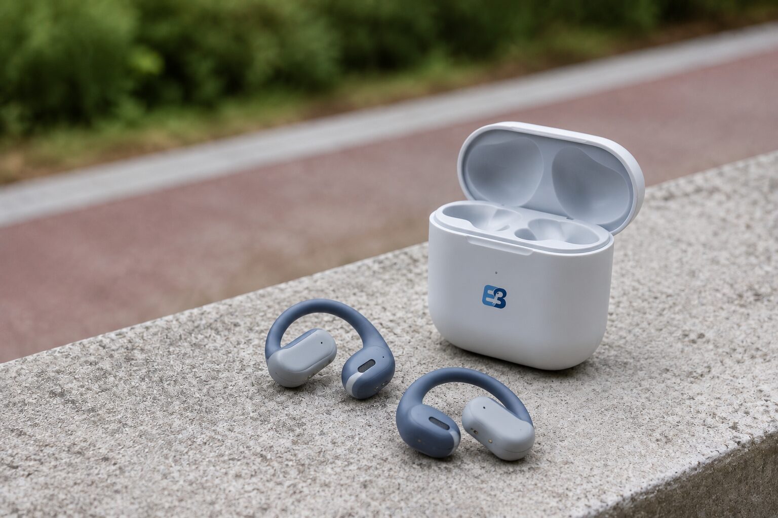 Open-ear hook earbuds with an EarbudSource branded charging case beside an outdoor running path